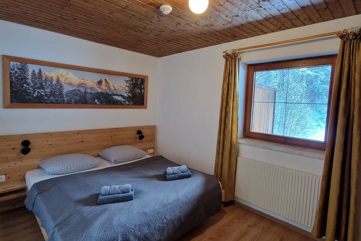Une chambre avec un plafond en bois, un lit recouvert d’une couverture grise, un tableau représentant un paysage de montagne et une fenêtre avec des rideaux.