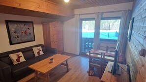 Een gezellige kamer met een houten plafond, een zwarte bank met rendierkussens, een houten salontafel en een zithoek met stoelen en een tafel.