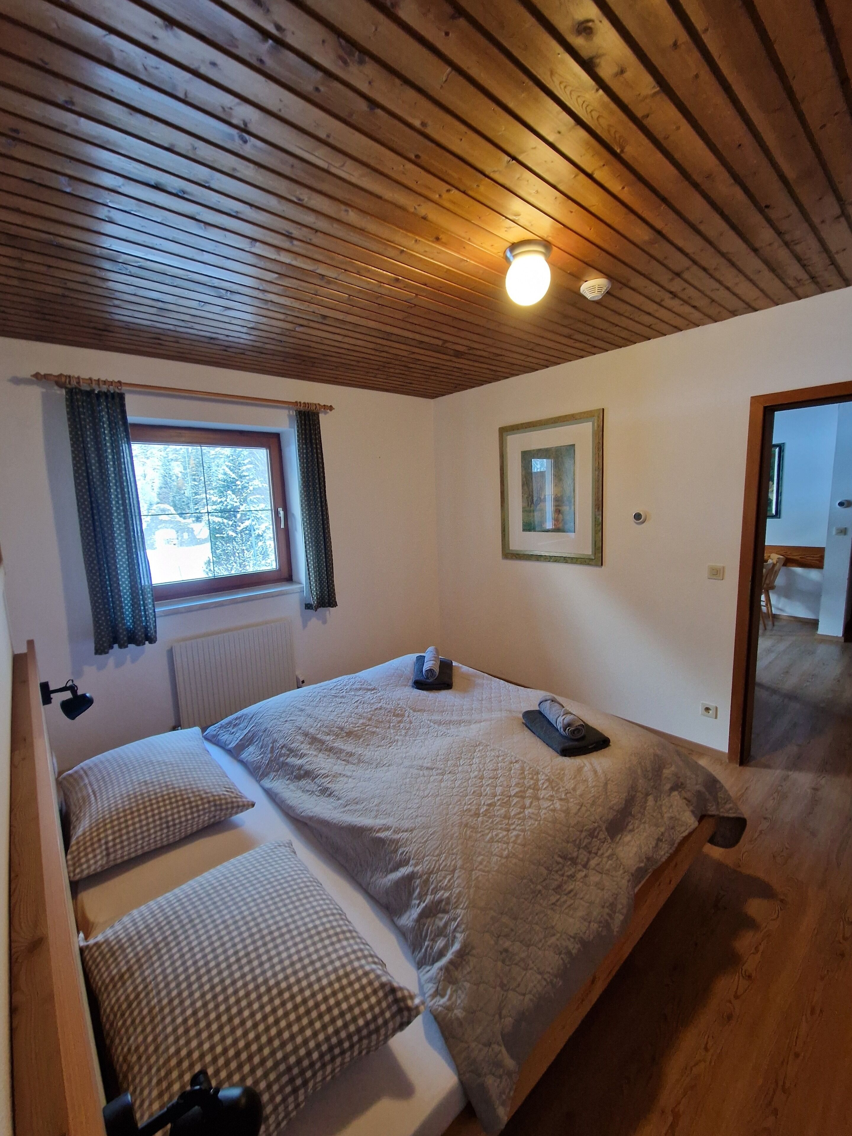 Un dormitorio con techo de madera, una cama con colcha, una ventana con cortinas e imagen enmarcada en la pared.