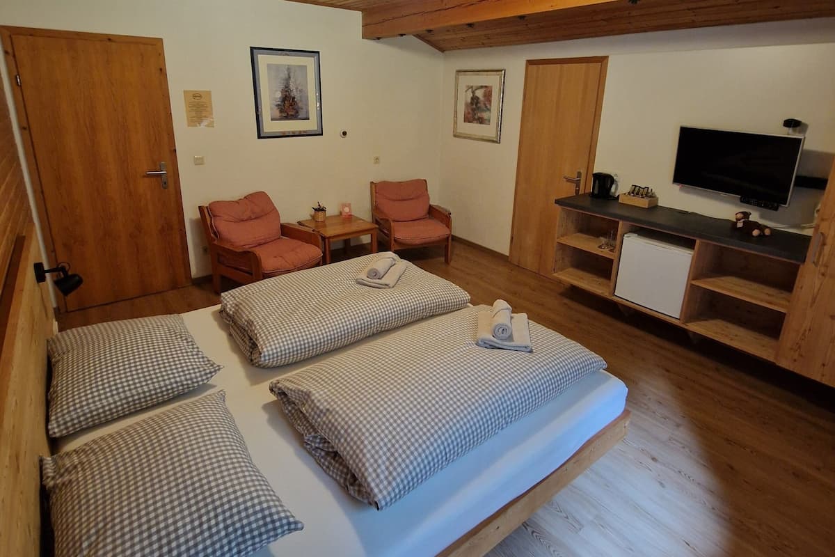 Une pièce chaleureuse avec un plafond en bois, un lit recouvert d’une couette à carreaux, deux chaises, une petite table, un téléviseur et un meuble blanc.
