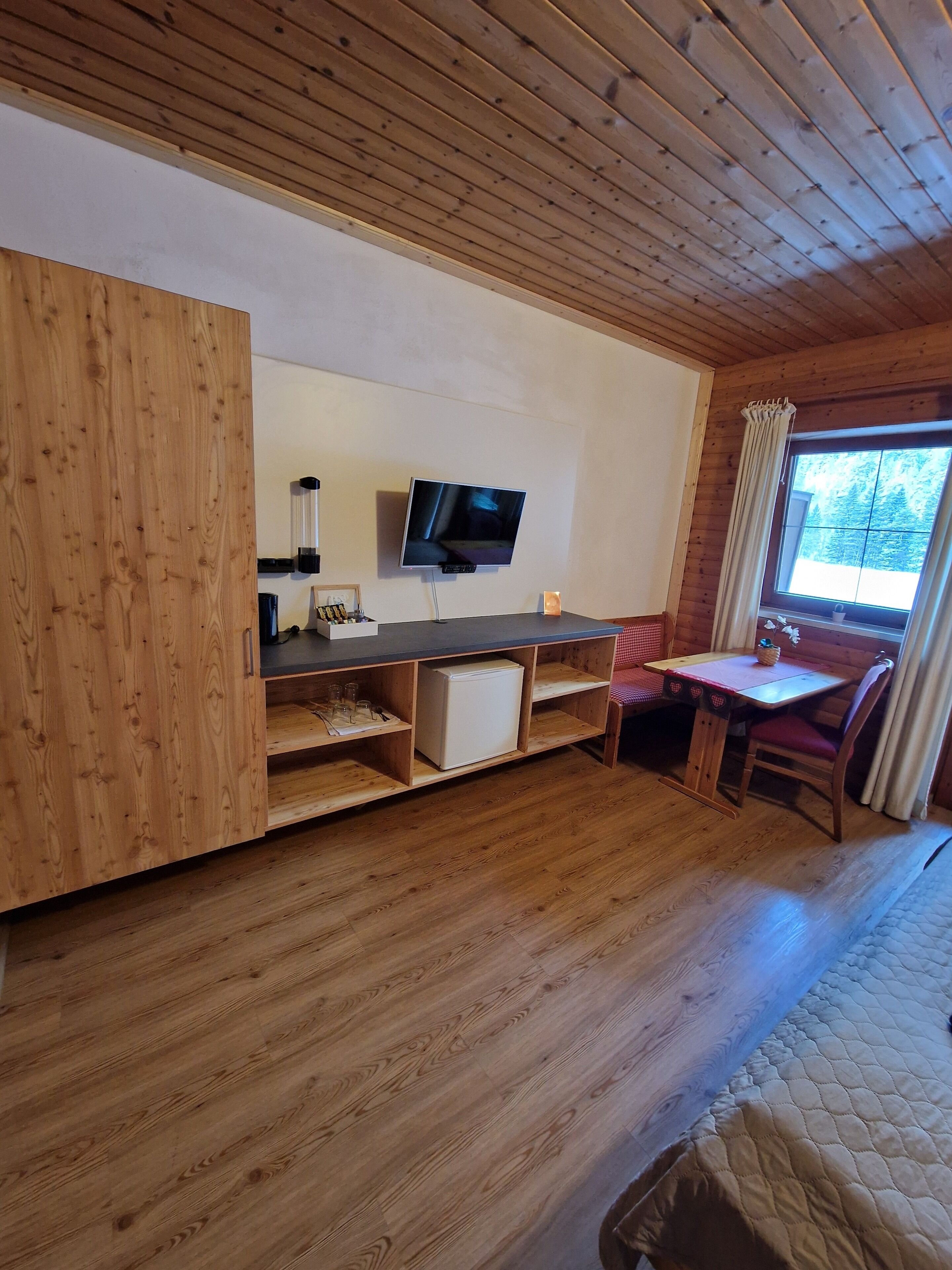 A wooden cabin interior with a TV mounted on the wall, a small table with a chair, and a window with curtains.