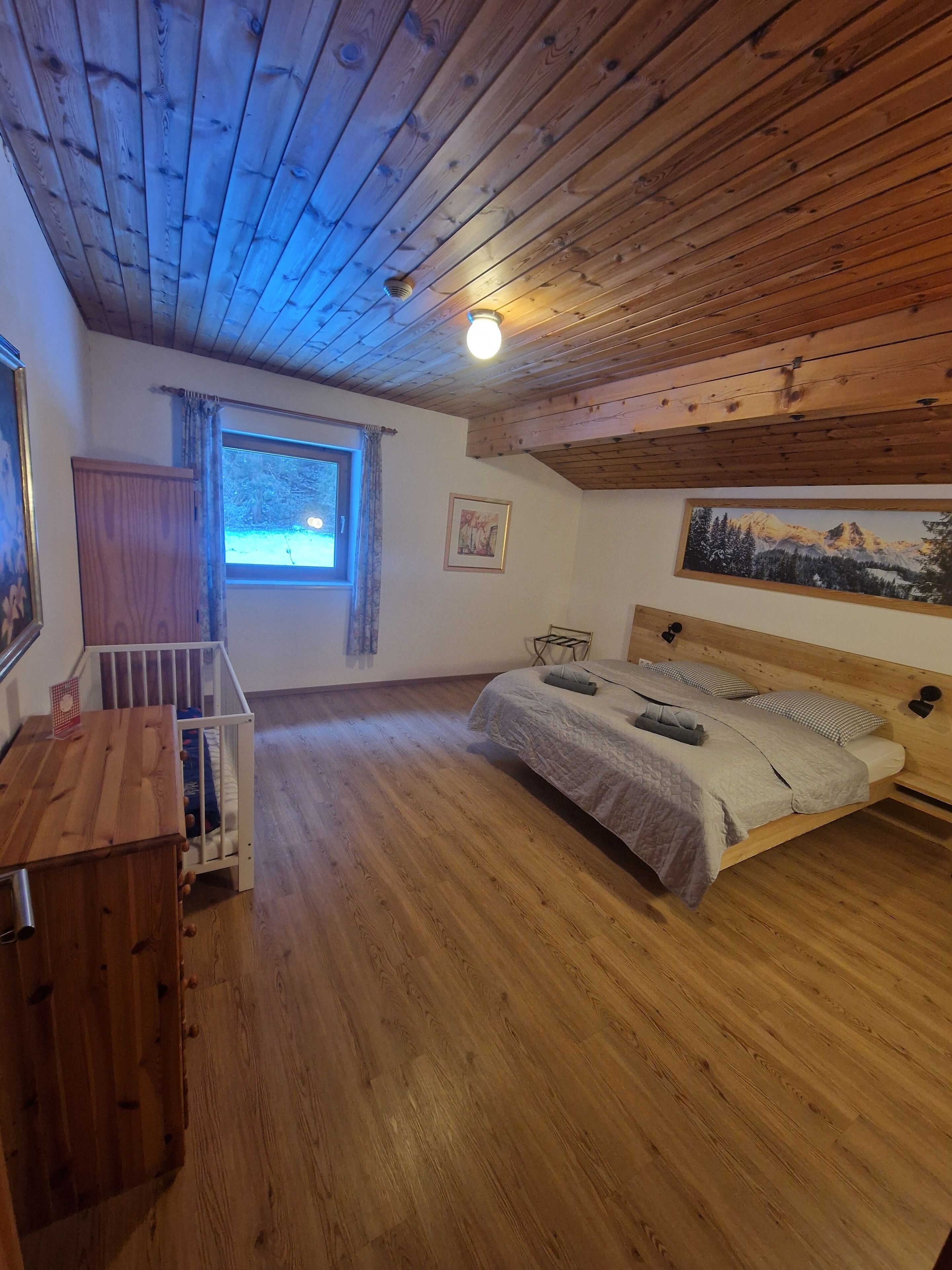 Ein gemütliches Zimmer mit Holzboden, einem Bett mit Kopfteil, einem Babybett, einem Fenster mit Vorhängen und einem Gemälde an der Wand.