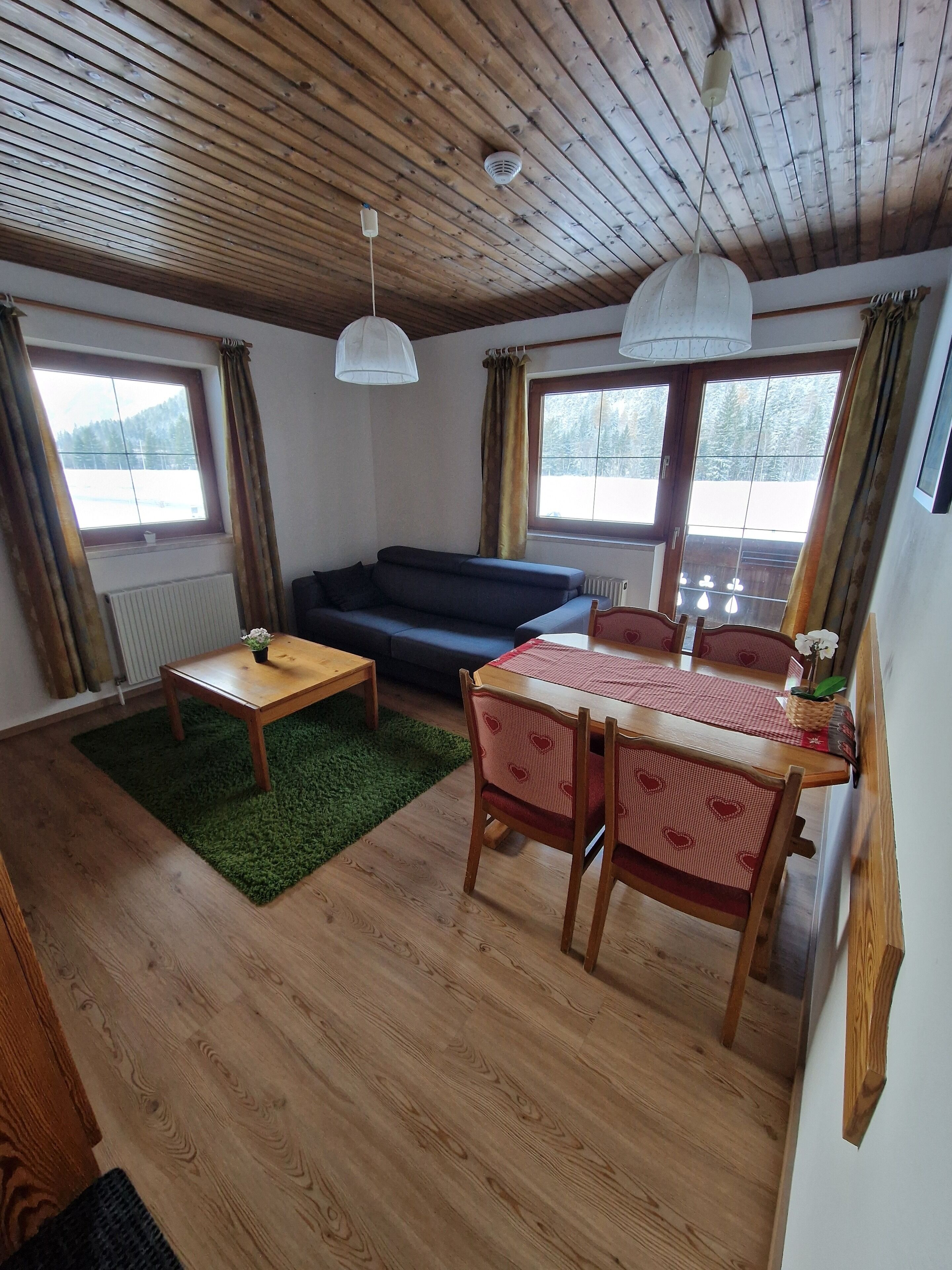A cozy living area with a wooden ceiling, a sofa, a dining table with chairs, and a view of a snowy landscape through large windows.
