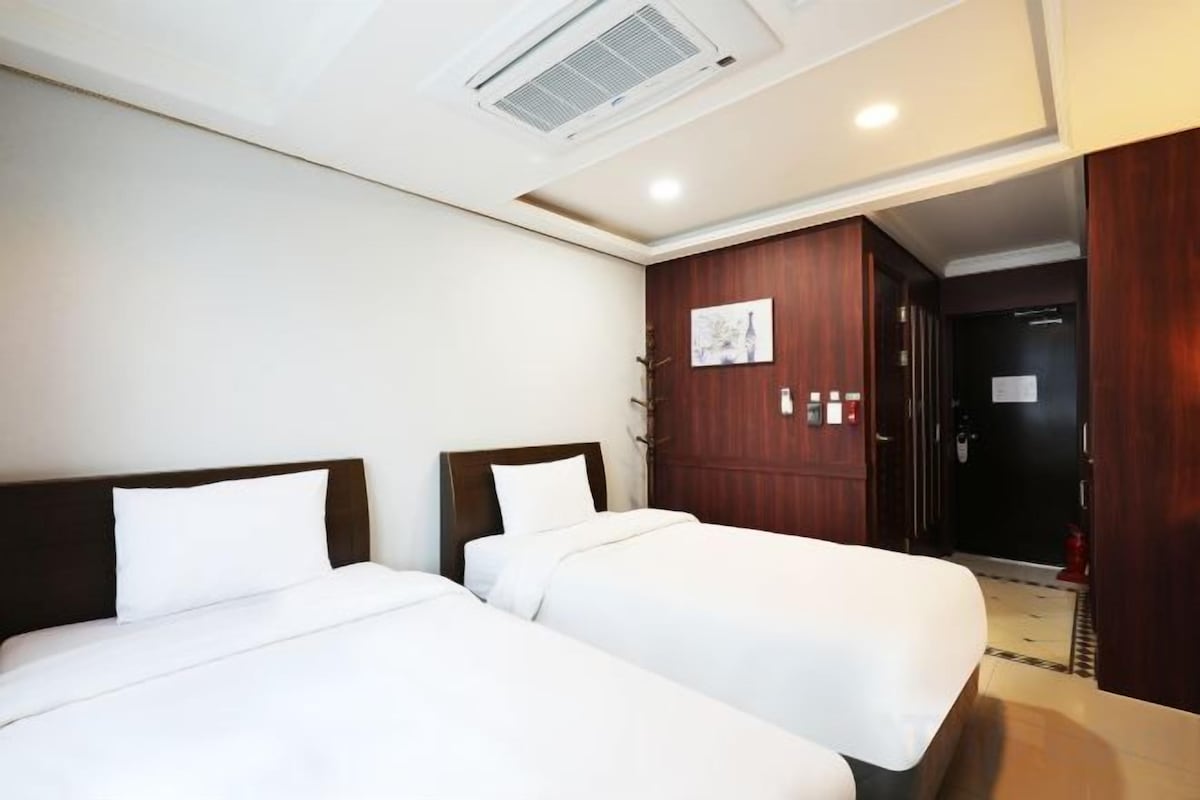 A hotel room with two beds, a wooden wardrobe, a ceiling vent, and a door leading to a hallway.