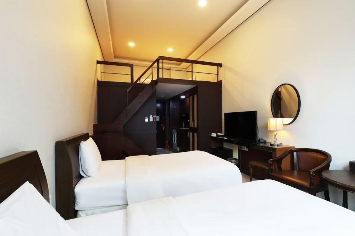 A hotel room with two beds, a desk, a chair, a TV, and a staircase leading to an upper level.