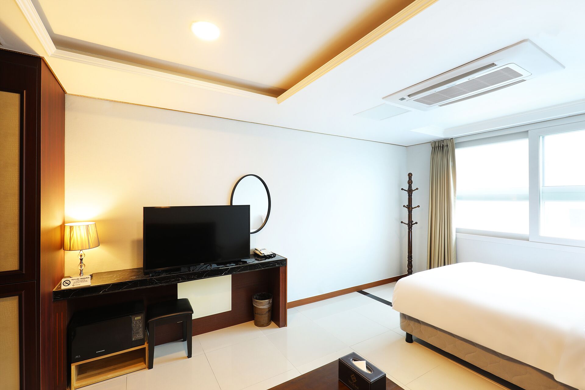 A hotel room with a bed, a desk with a television, a lamp, a coat rack, and a window with curtains.