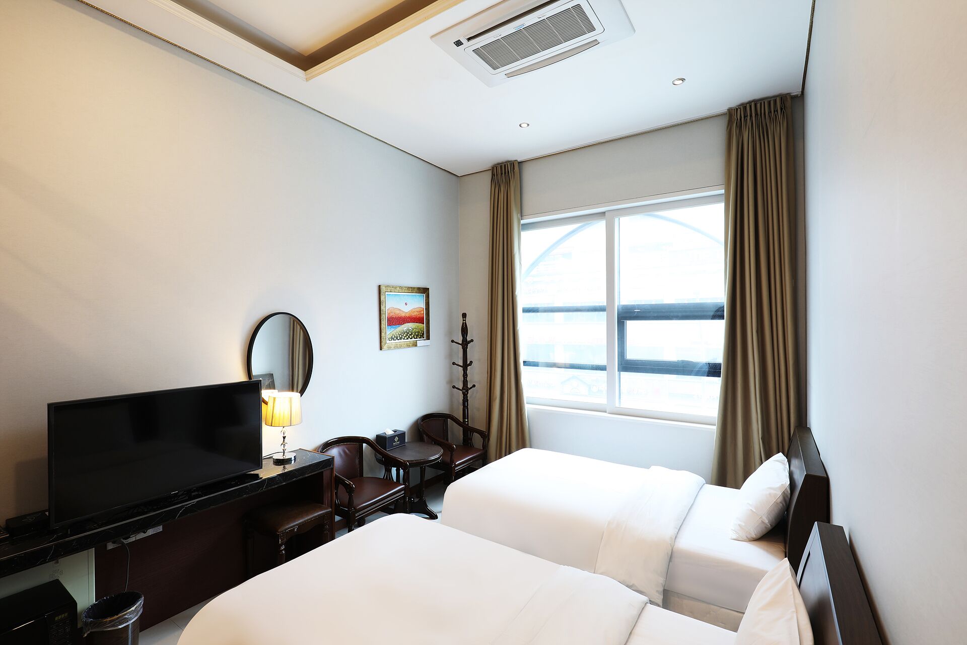 A hotel room with a flat-screen TV, a desk, two beds, a chair, a coat rack, a window with curtains, and a painting on the wall.
