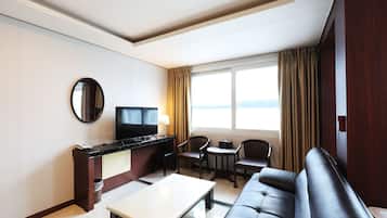 A hotel room with a flat-screen TV, a sofa, a coffee table, two armchairs, a small round table, and a large window with curtains.