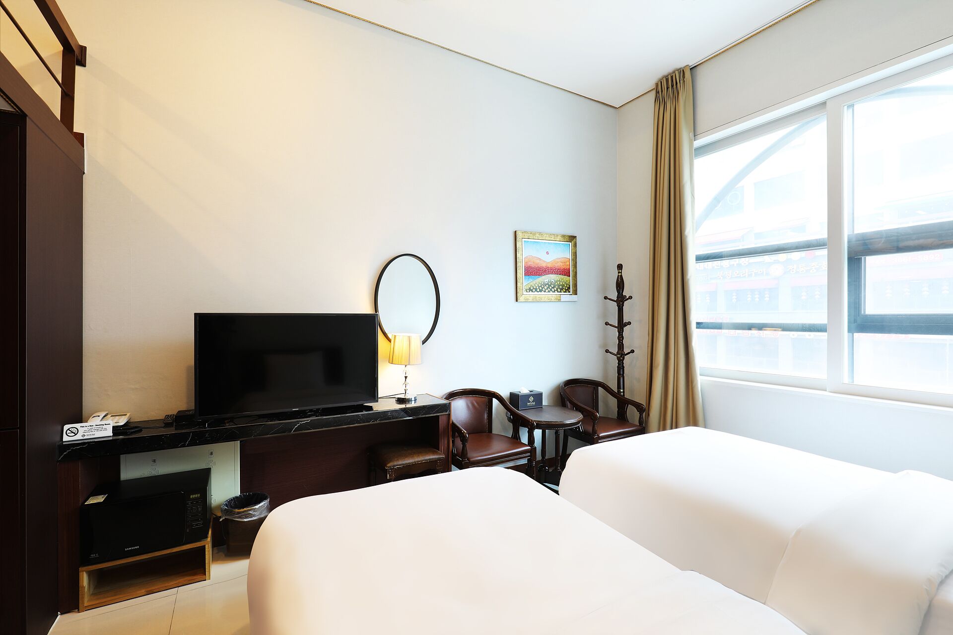 A hotel room with a flat-screen TV, a desk with a lamp, two chairs, a bed, and a window with curtains.