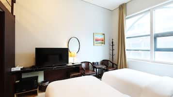 A hotel room with a flat-screen TV, a desk with a lamp, two chairs, a bed, and a window with curtains.