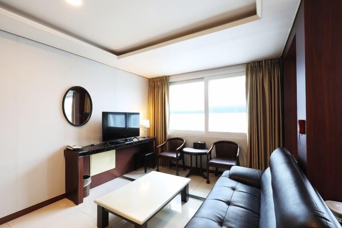A hotel room with a sofa, a small dining table, a TV, and a window with curtains.