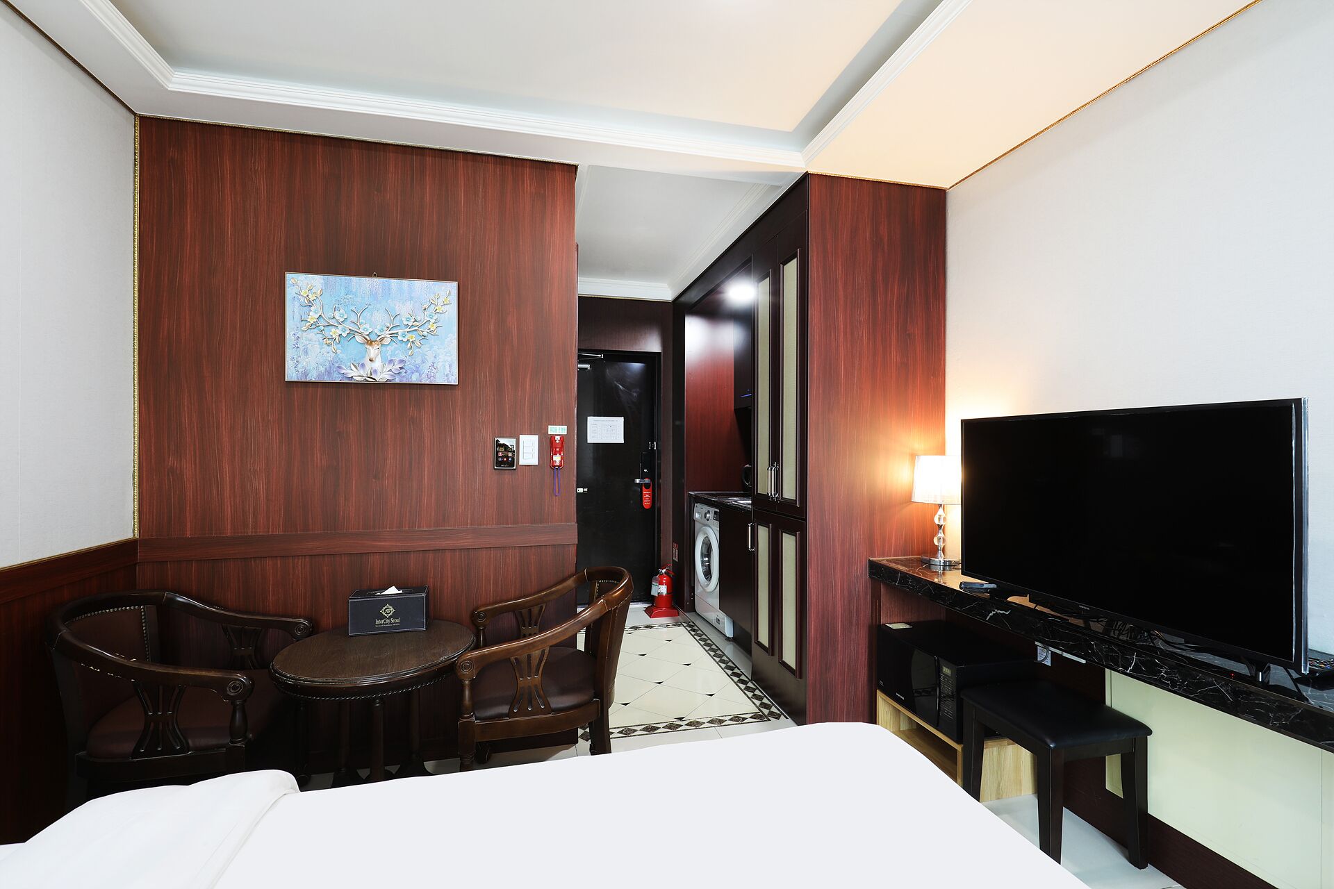 A hotel room with a bed, a desk with a chair, a TV, a washing machine, and a painting on the wall.