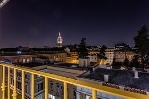 A nighttime cityscape with a prominent tower in the distance, surrounded by buildings with balconies and a yellow railing in the foreground.