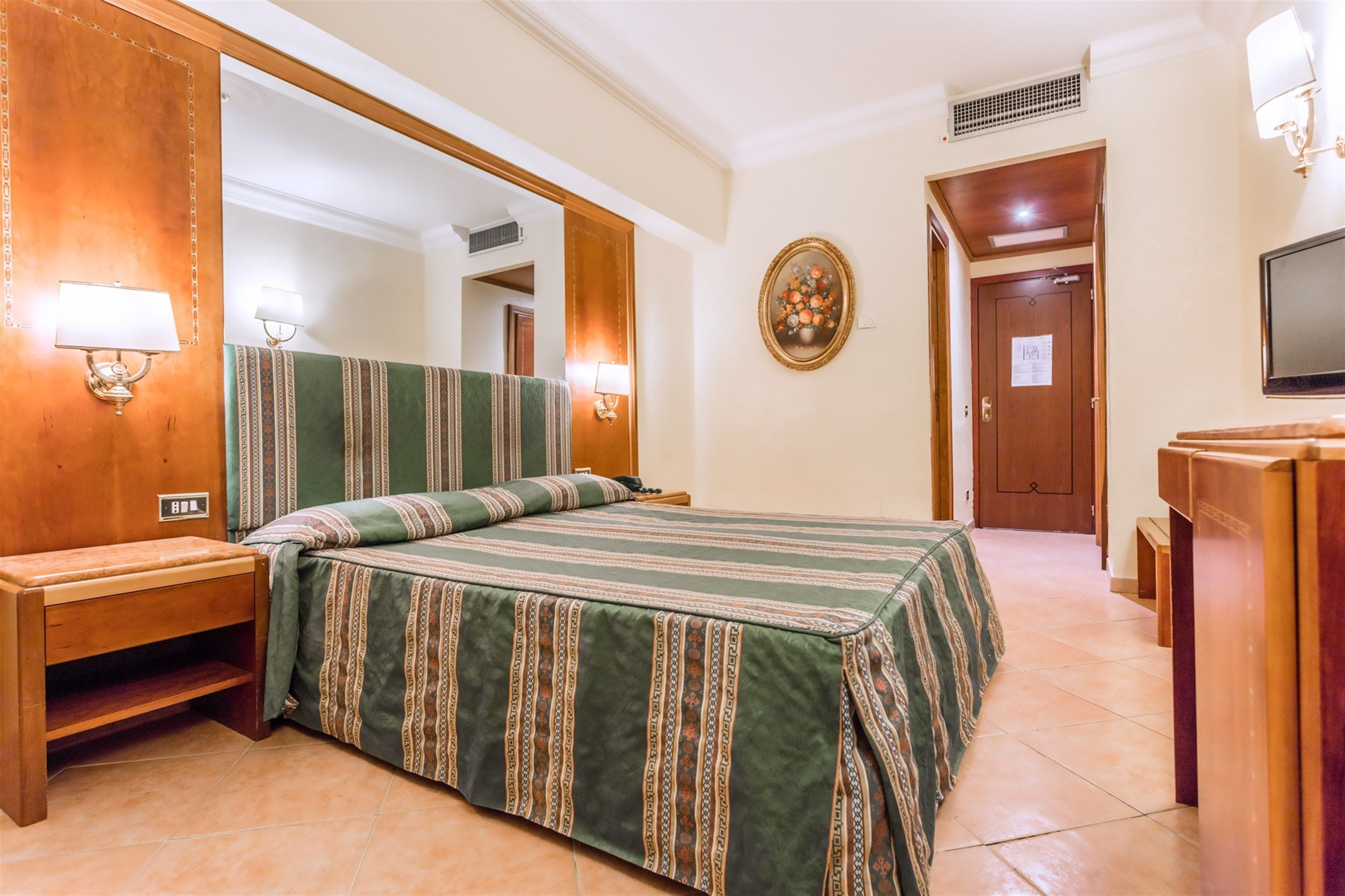A hotel room with a large bed, wooden headboard, bedside tables, a mirror, a television, and a door with a sign.
