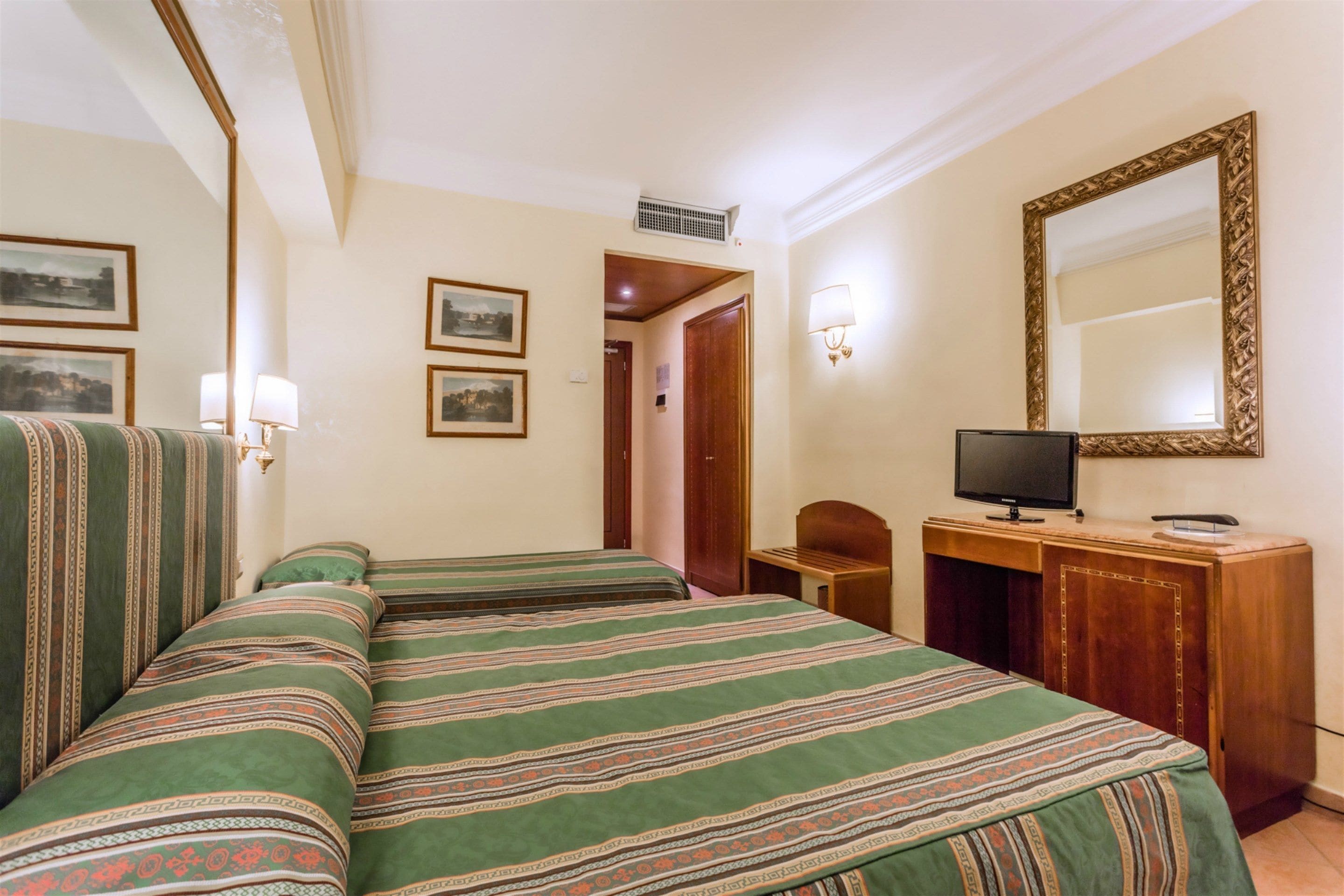 Quarto de hotel com duas camas, uma escrivaninha, televisão e quadros na parede.