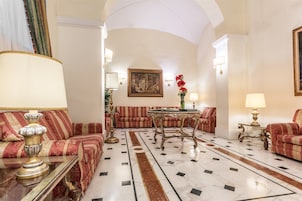 A spacious lobby with a high ceiling, red upholstered sofas, a marble floor, and a central table with a vase of flowers.