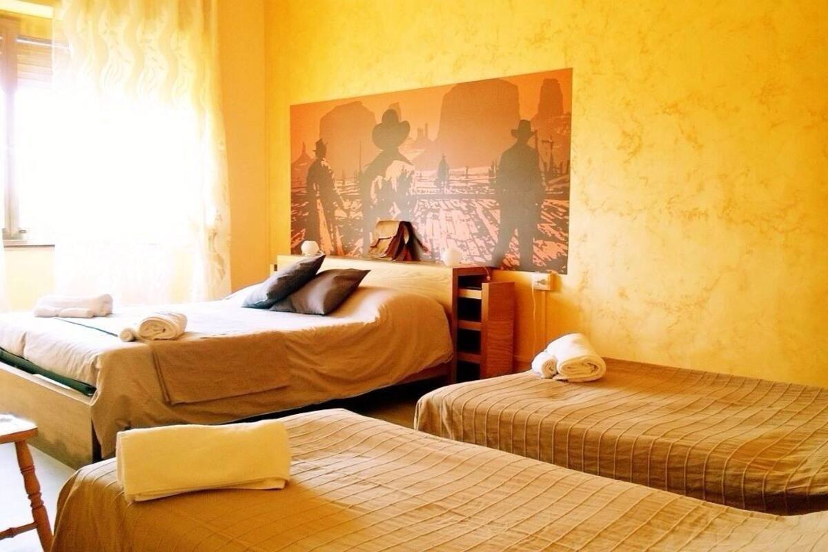 A hotel room with two beds, each with a pillow and a rolled towel. There is a large painting on the wall depicting cowboys.