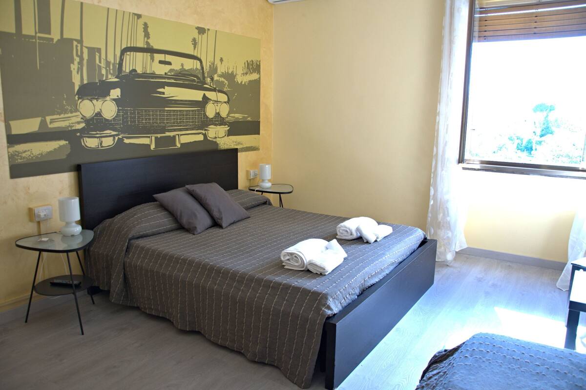 A hotel room with a bed, two bedside tables, a lamp, a painting on the wall, and a window with curtains.