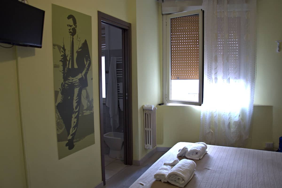 A hotel room with a bed, towels, a radiator, a window with blinds, a toilet, and a television on the wall.