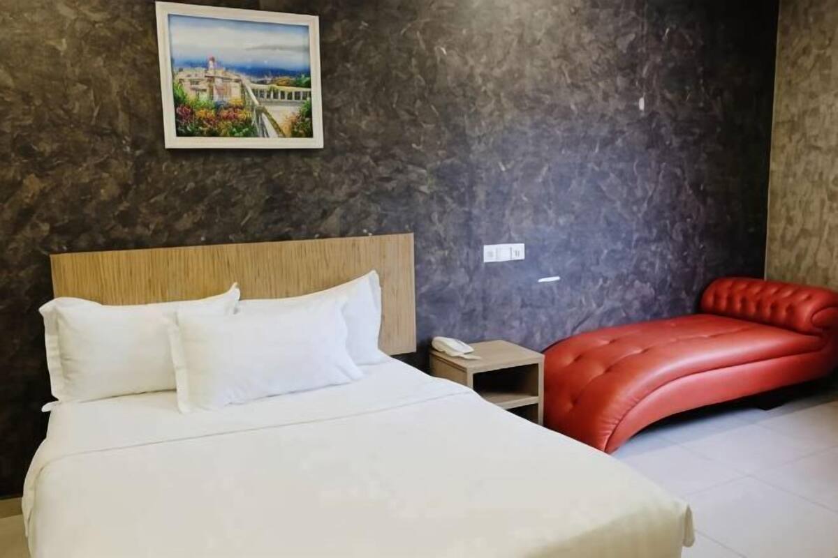A hotel room with a bed, a red leather sofa, a nightstand, and a painting on the wall.