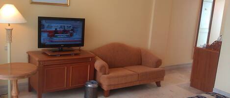 A living room with a television on a wooden cabinet, a small round table, a sofa, and a trash can.