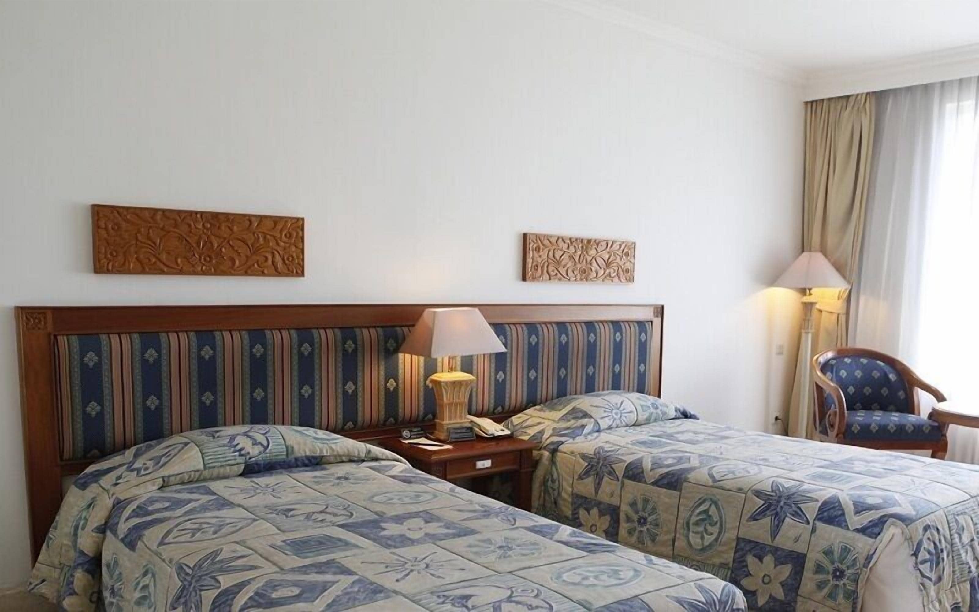 A hotel room with two beds, a wooden headboard, decorative wall art, a nightstand with a lamp, and a chair.