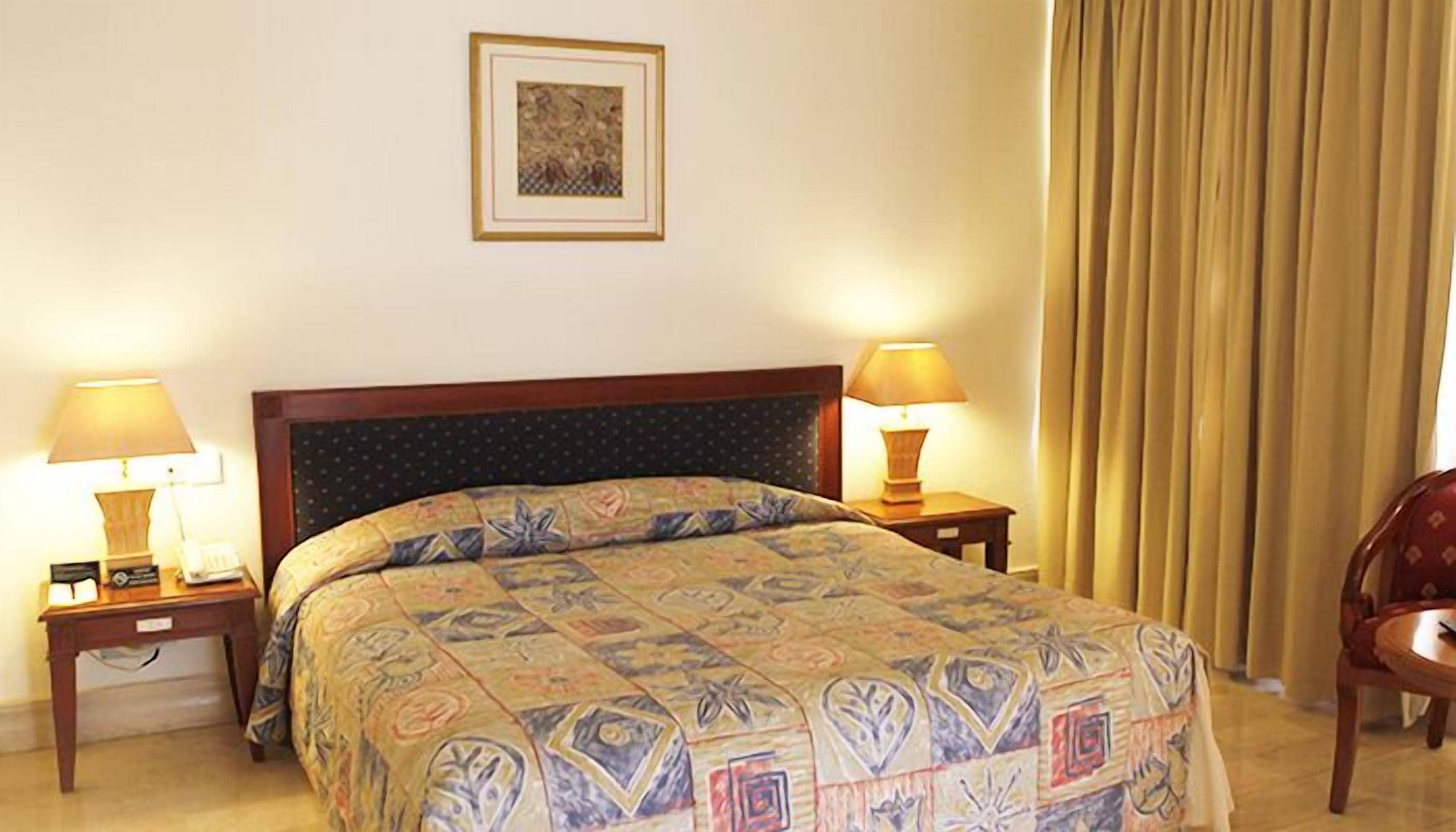 A hotel room with a bed, two bedside tables with lamps, a framed picture on the wall, and a chair by the window.