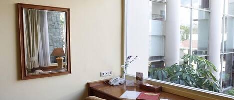 A hotel room with a wooden desk, a telephone, a lamp, and a mirror. There is a window with a view of a building and a plant outside.