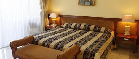 A hotel room with a large bed, a bench, two bedside lamps, a framed picture on the wall, and a window with curtains.
