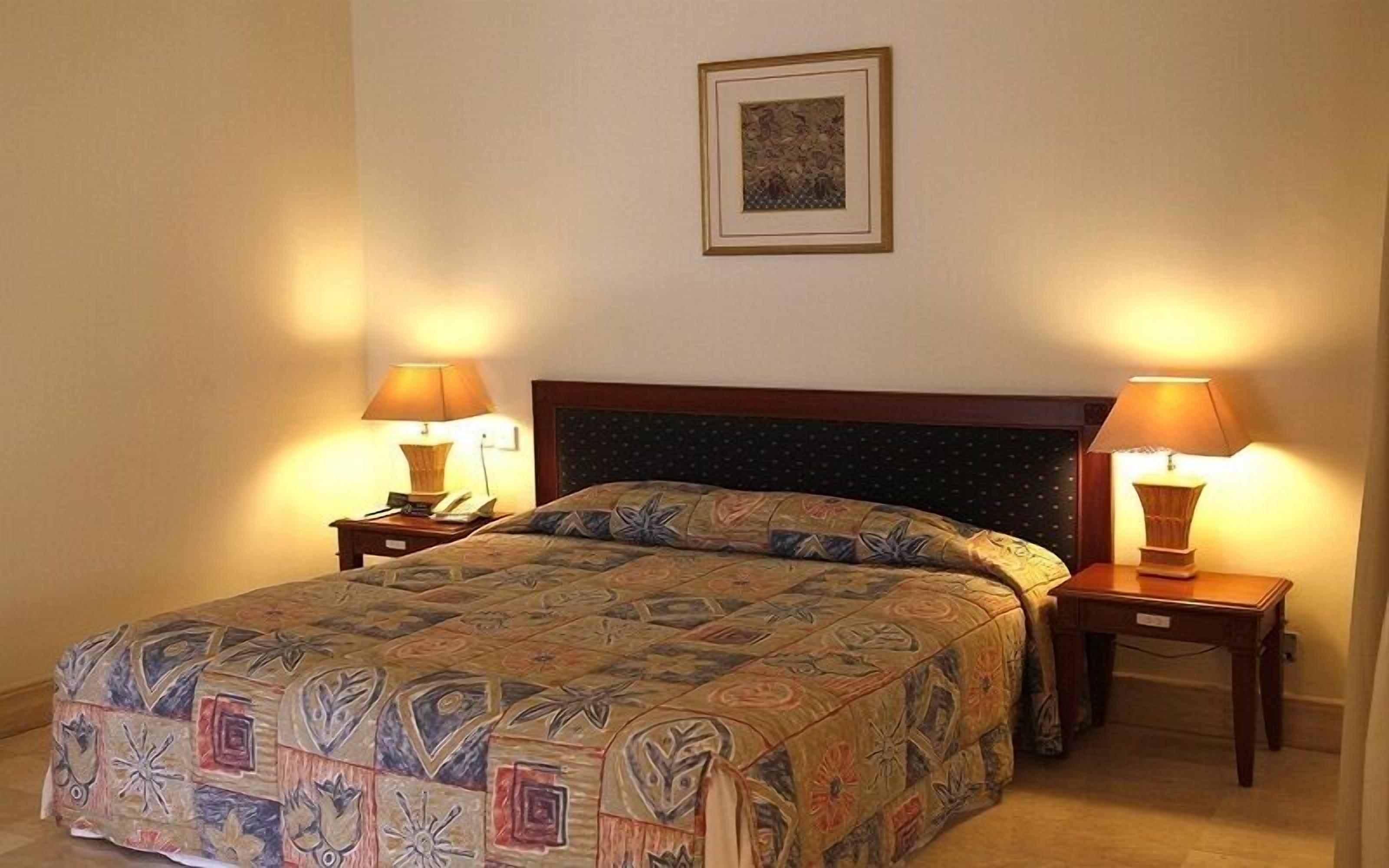 A double bed with a patterned bedspread, two bedside lamps, a framed picture on the wall, and a wooden headboard.
