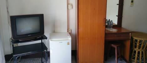 Una habitación pequeña con televisión sobre un soporte, un refrigerador compacto, un escritorio de madera con espejo y un taburete de mimbre.