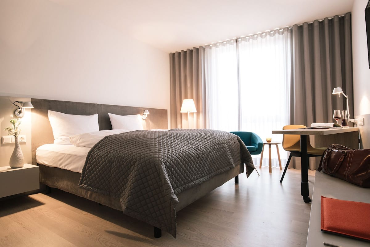Una habitación de hotel con cama, lámparas de noche, escritorio, silla y ventana con cortinas.