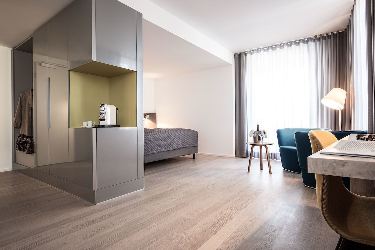 Una habitación de hotel moderna con una cama, una zona de estar con una mesa y sillas, una cafetera y un armario.