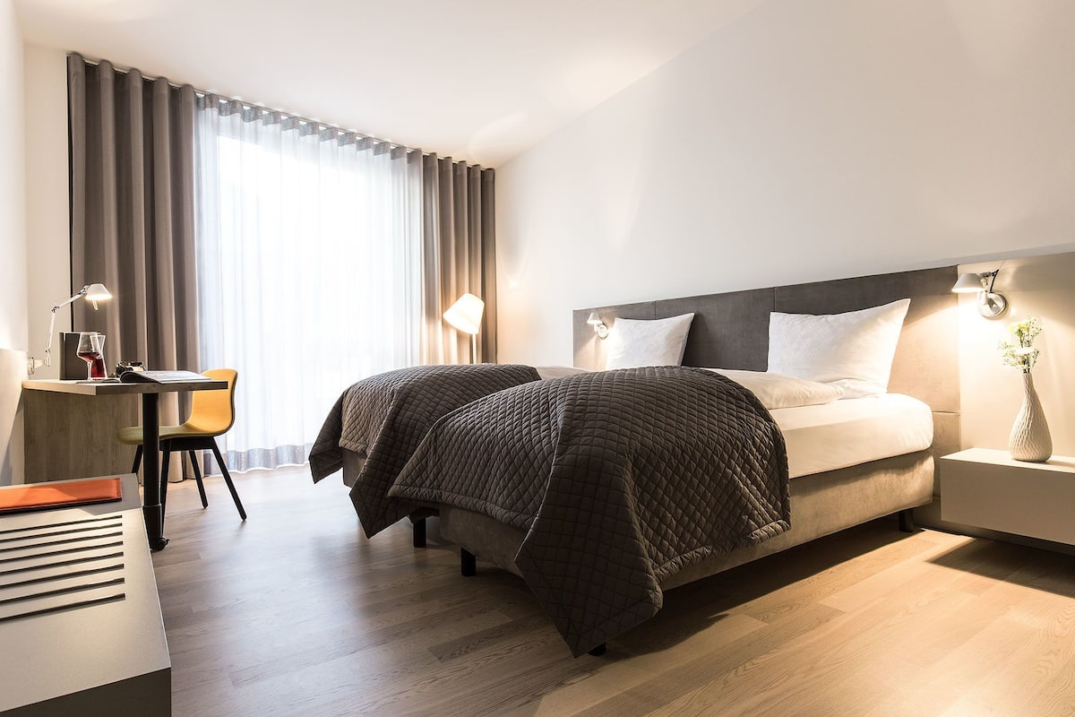 Una habitación de hotel con cama, mesita de noche, lámpara, silla, escritorio y cortinas.