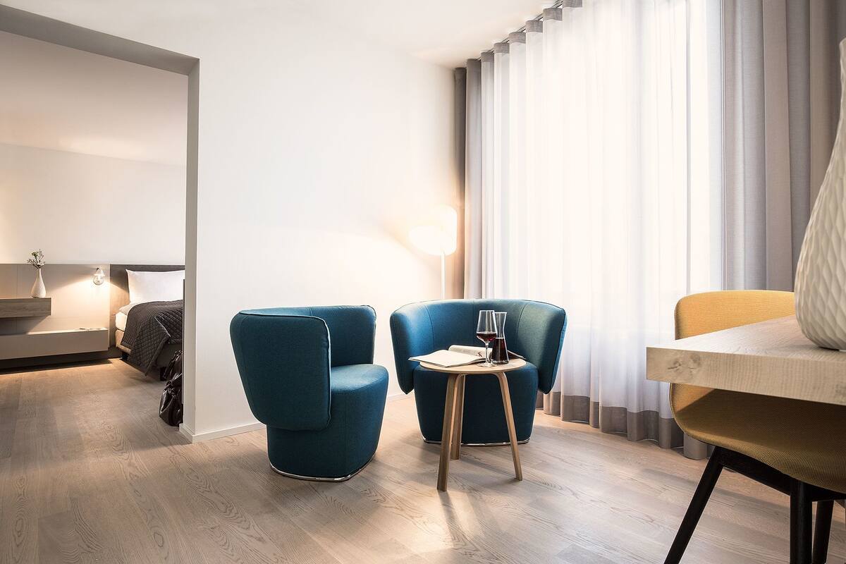 Una habitación de hotel moderna con una cama, dos sillones azules, una mesita redonda con una copa de vino y una lámpara.