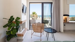 A modern living room with a large green sofa, a wicker chair, and a small table. A potted plant is on the left, and a TV is mounted on the wall. The room has a view of the beach and ocean through large glass doors.