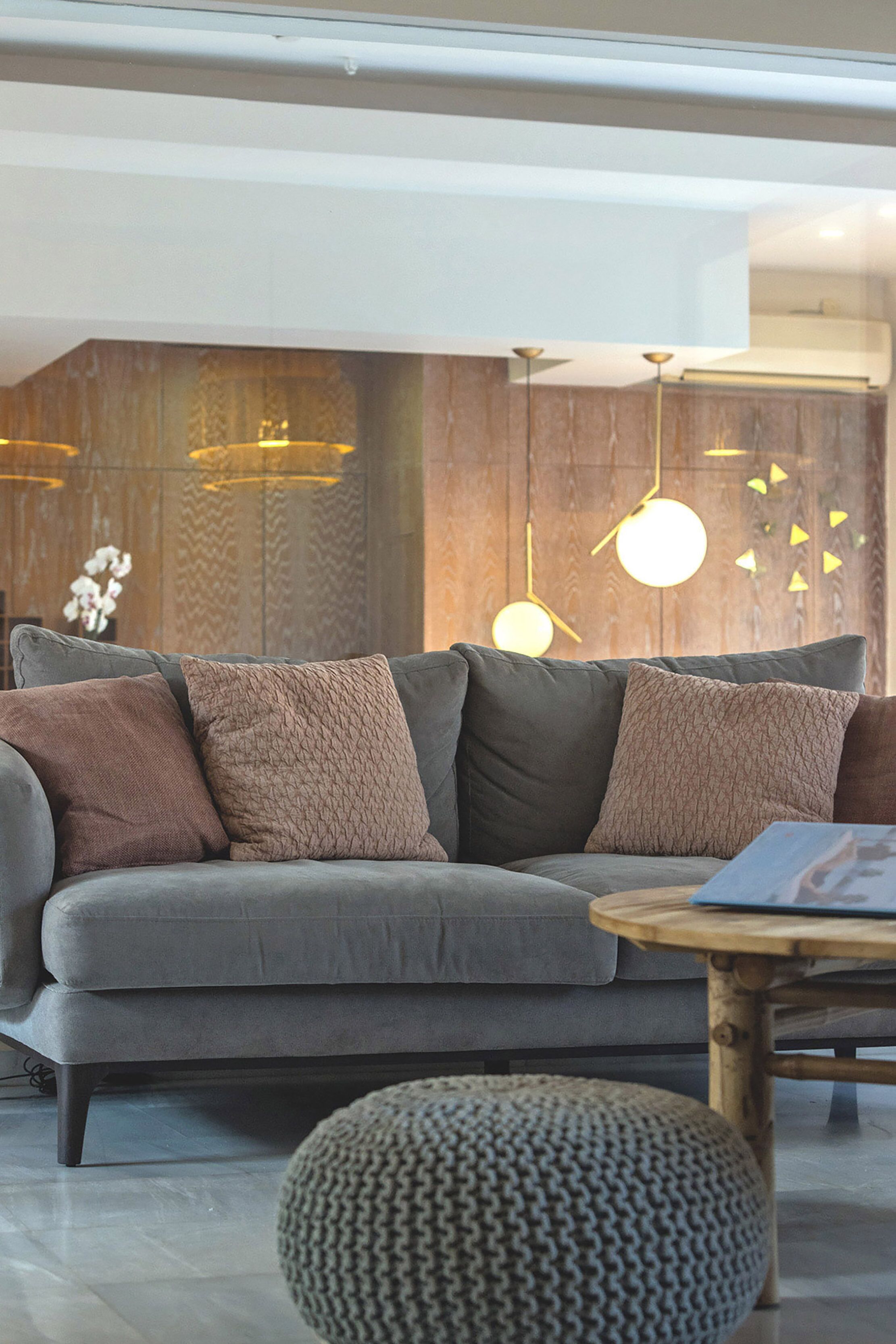 Una sala de estar moderna con un sofá gris, varios cojines, una mesa de centro redonda de madera y un pouf tejido.
