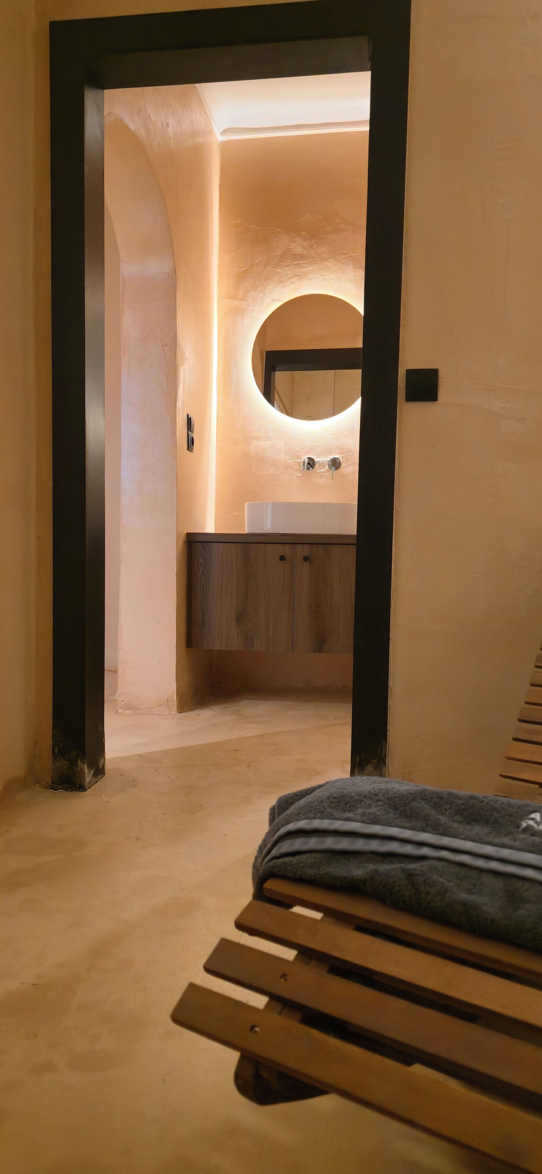A hallway leading to a bathroom with a wooden bench, a round mirror, and a sink.