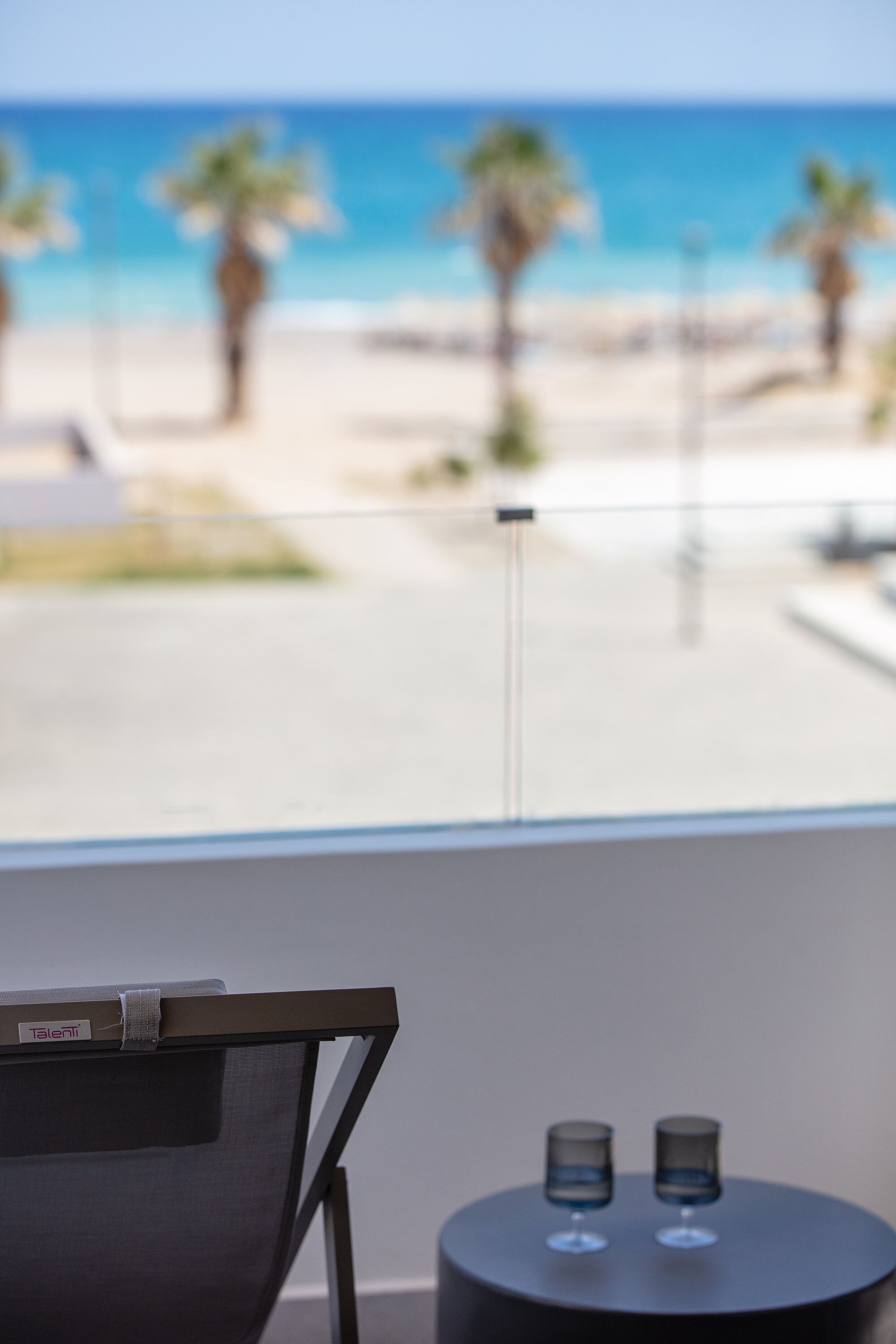 A room with a view of the beach, a chair, and a small table with two glasses.
