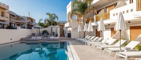 A swimming pool with lounge chairs and umbrellas, surrounded by a building with balconies and a clear sky.