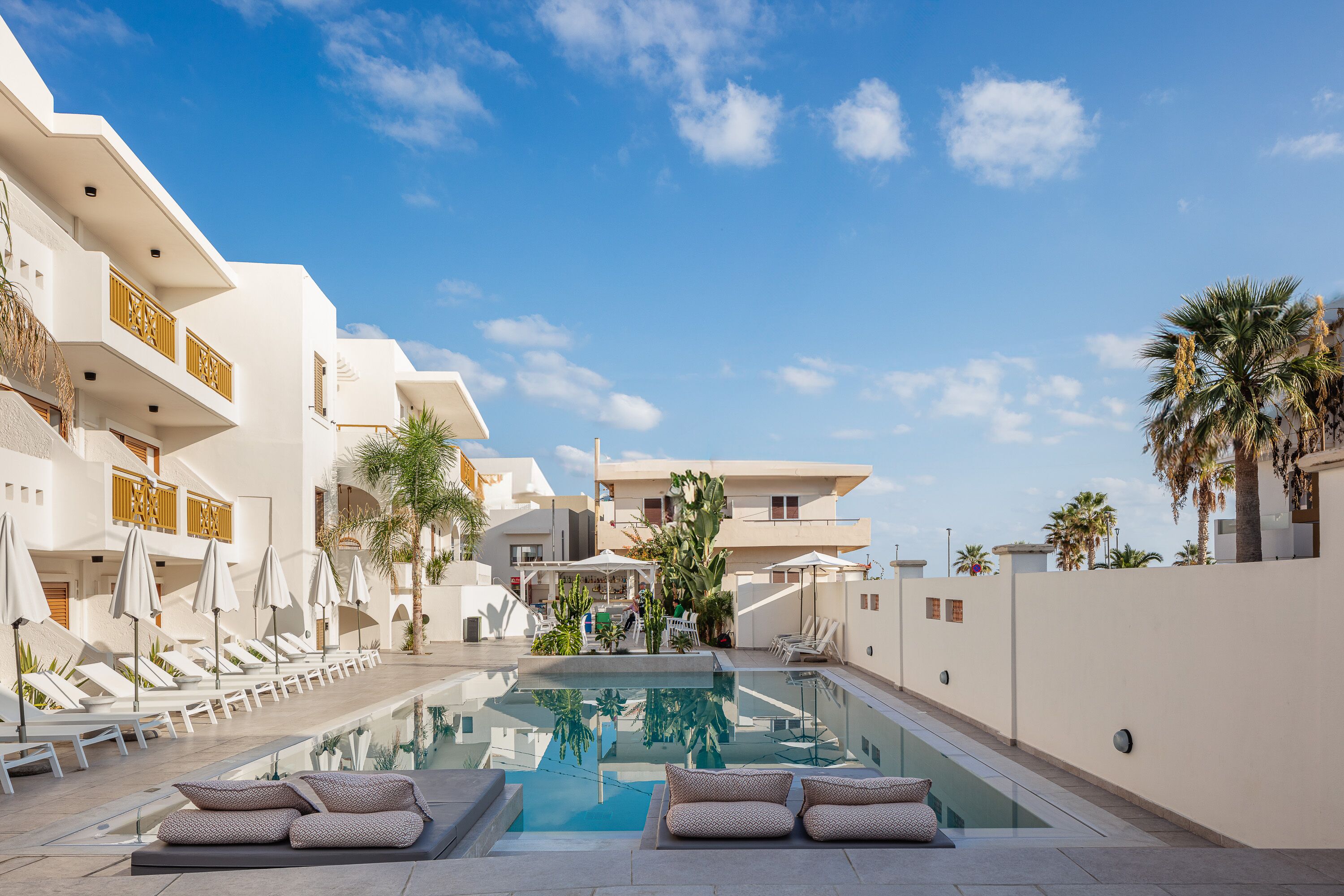 Un área de piscina con sillones de descanso y sombrillas, rodeada de edificios blancos con balcones y palmeras.