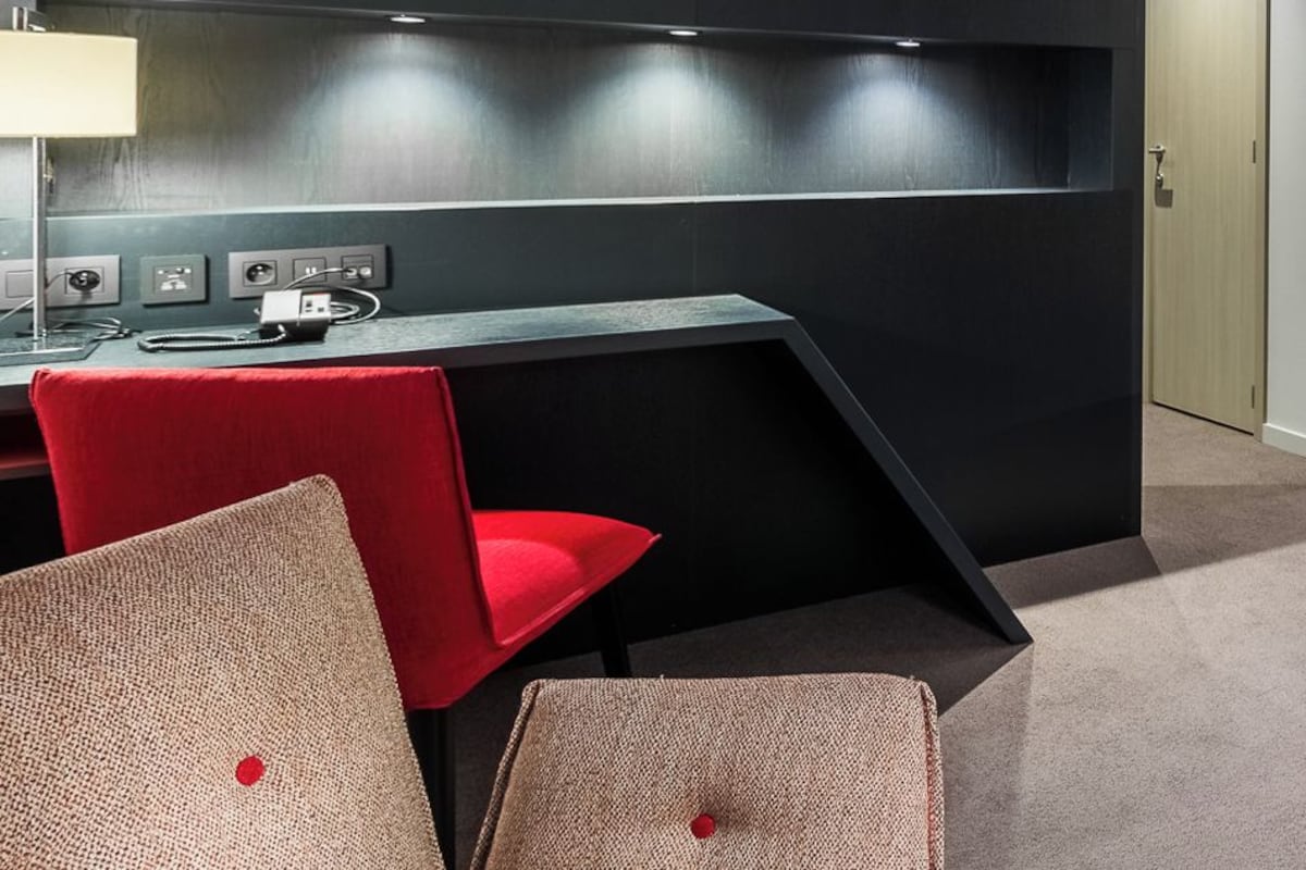 A hotel room with a desk, two chairs, a bed with red bedding, and a modern design.