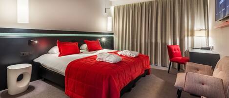 Un cuarto de hotel con una cama grande, almohadas rojas, una colcha roja, una silla moderna, una mesita pequeña y un televisor montado en la pared.