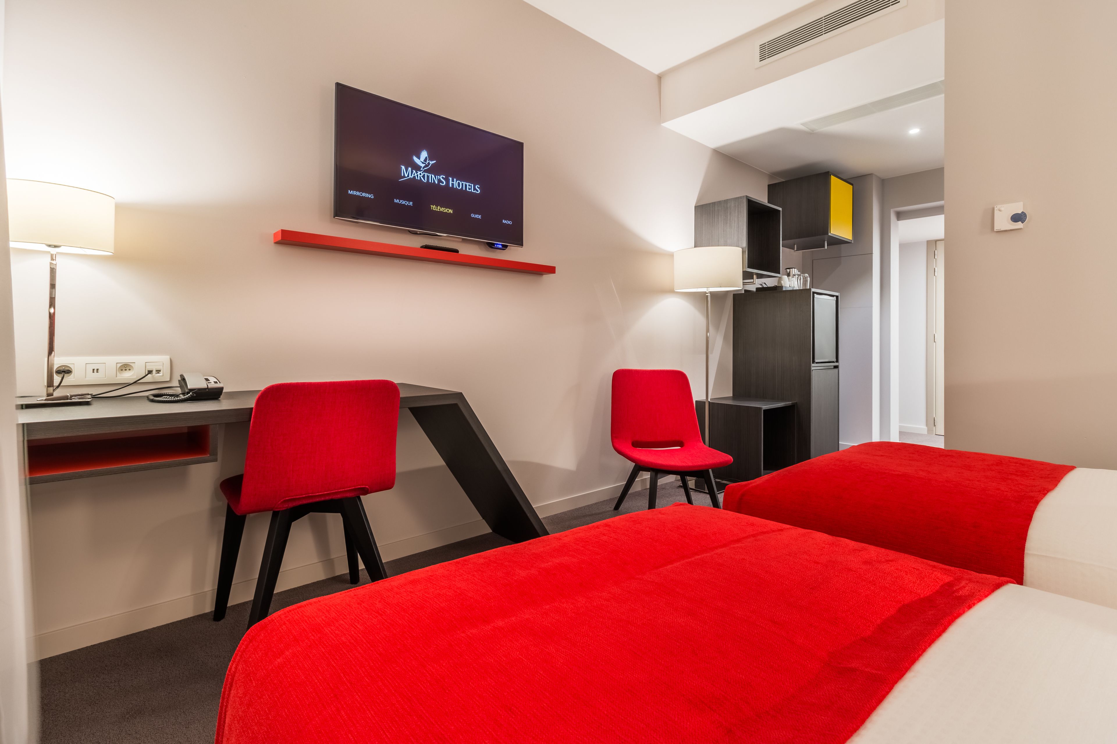 A hotel room with a TV displaying 'Martin's Hotels' and a desk with two red chairs.