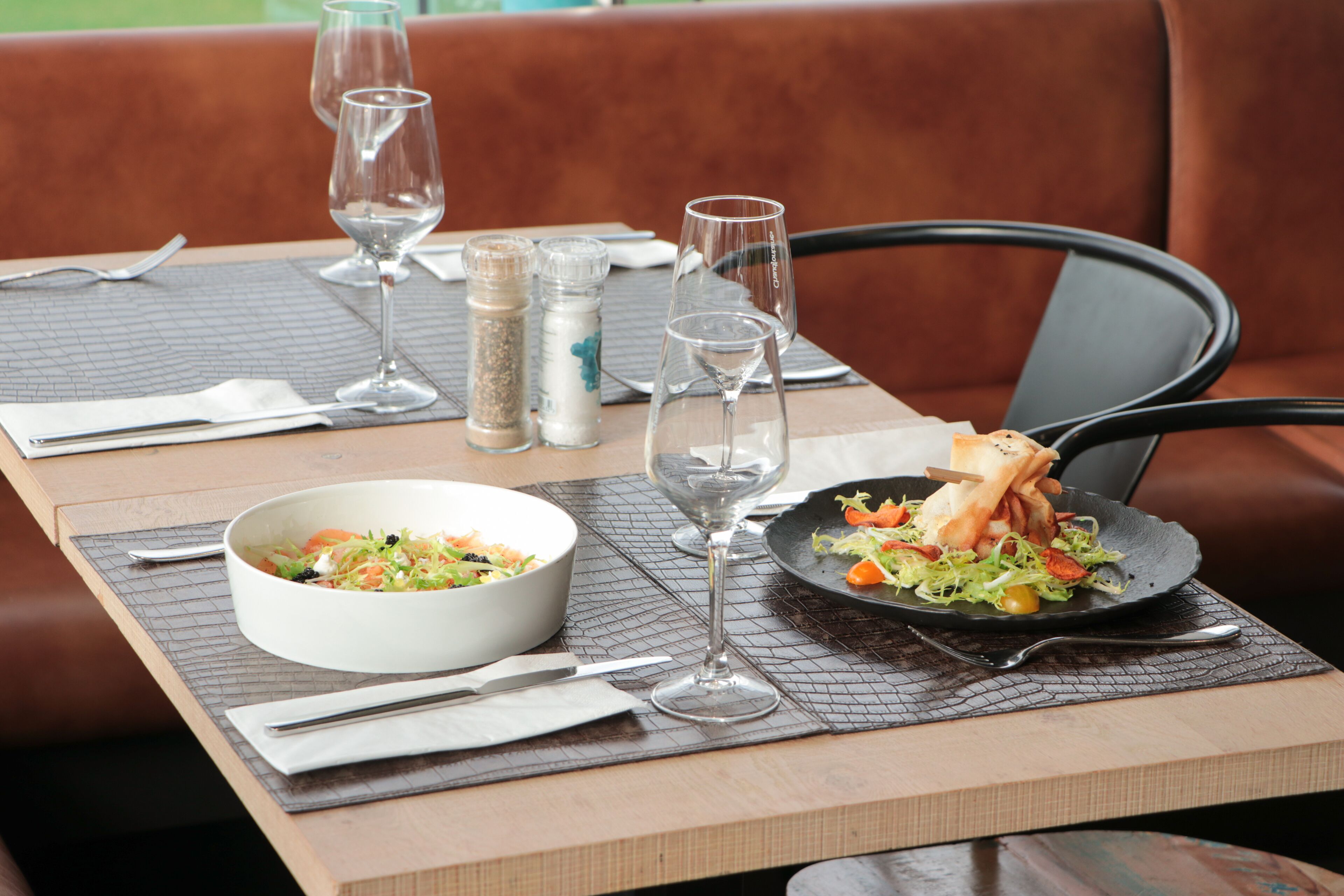 Una mesa de comedor con ensalada en un bol, un plato de comida, copas de vino y cubiertos.