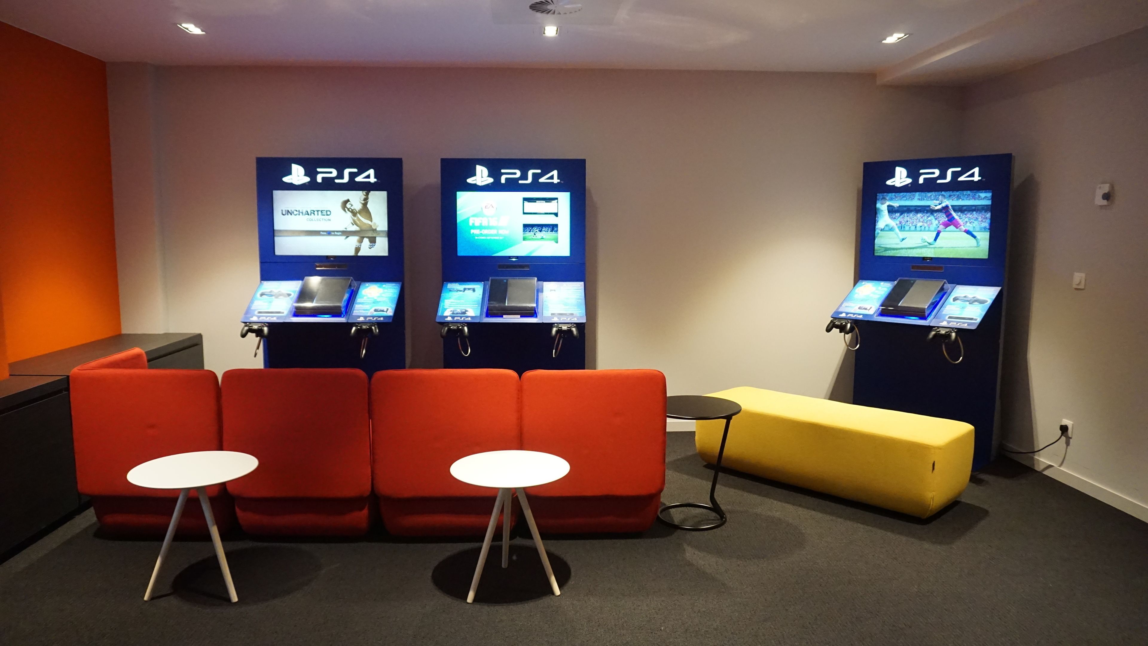 Et gaming lounge med tre PS4 spille stationer, røde siddepladser og en gul fodskammel.
