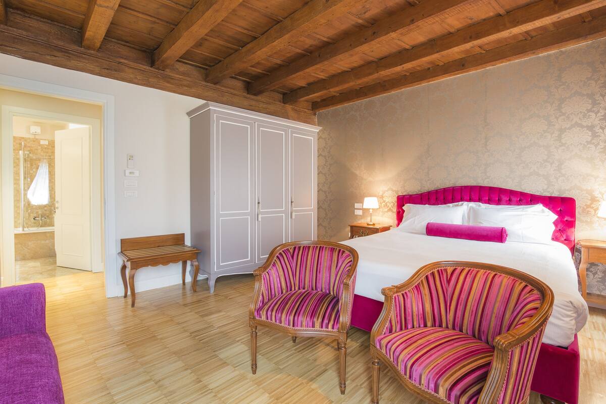 Un dormitorio con una cama grande, dos sillones a rayas, un banco de madera y un armario.