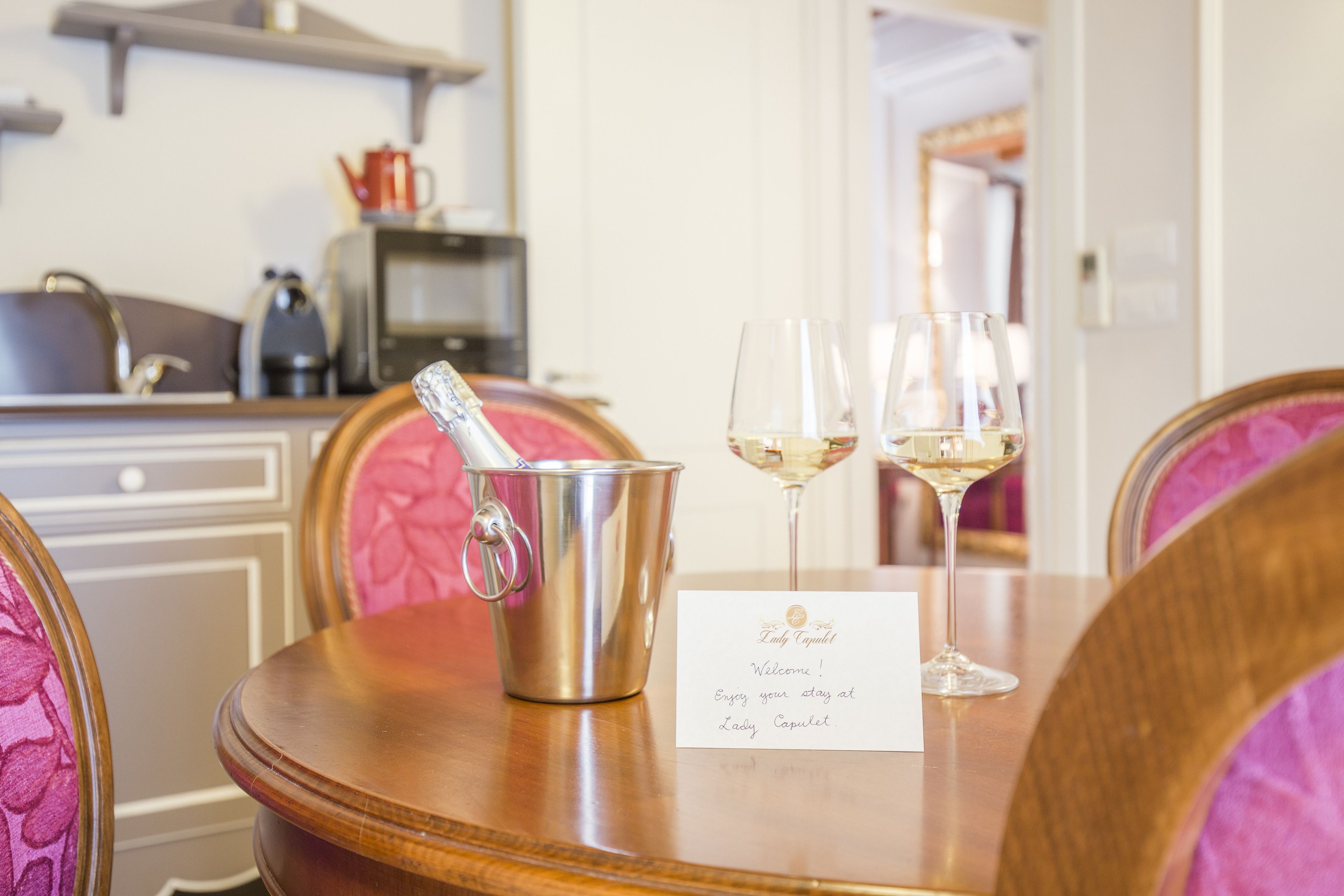 Una mesa con un cubo de champán, dos copas de vino y una tarjeta de bienvenida en una habitación con sillas tapizadas en rosa y una mesa de madera.