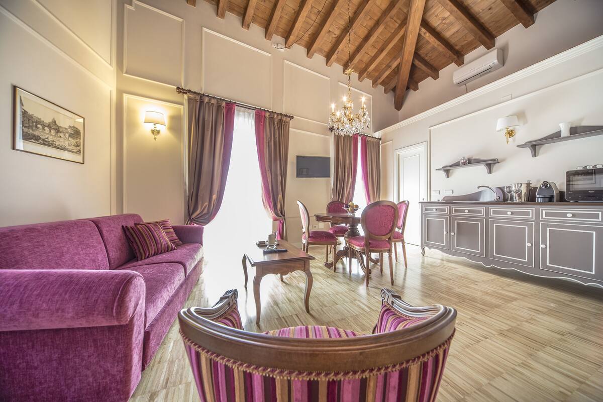 Una sala de estar con un sofá púrpura, un comedor con una araña, y una cocina con armarios de madera.
