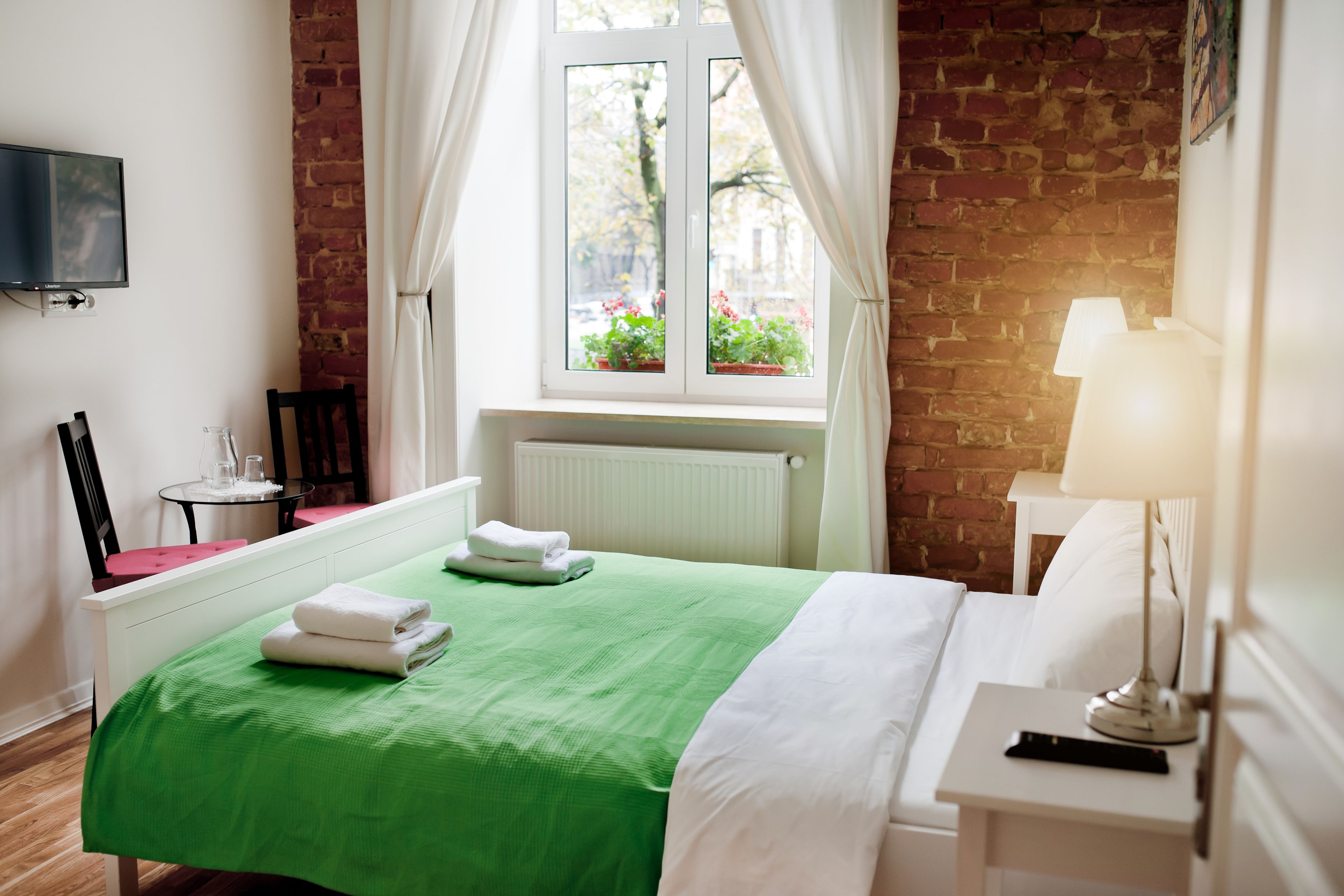 A hotel room with a green bedspread, white linens, a white nightstand with a lamp, a TV mounted on the wall, a brick wall, and a window with curtains.